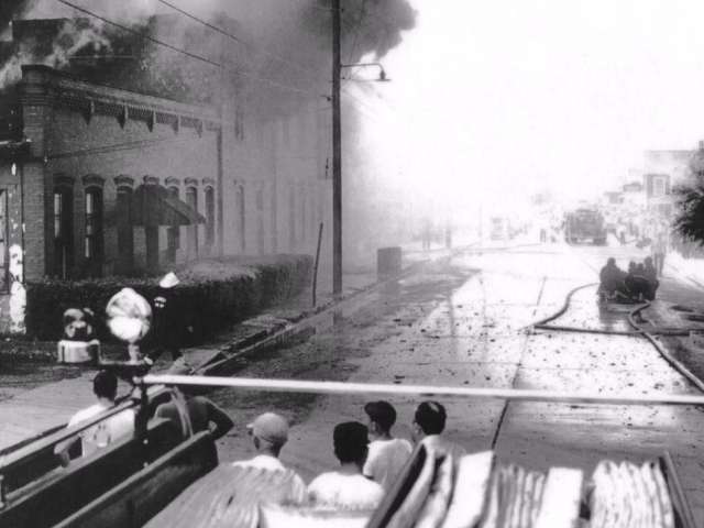 A vintage photo of a fire blazing in a building as people look on. 