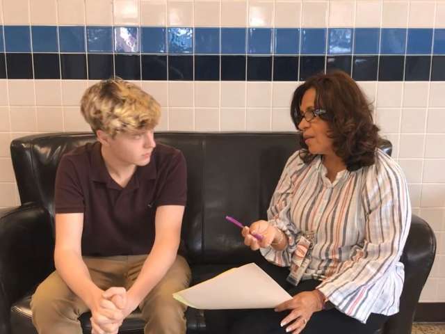 A young man with blonde hair sits next to a woman with a striped shirt on a leather sofa in a school hallway.