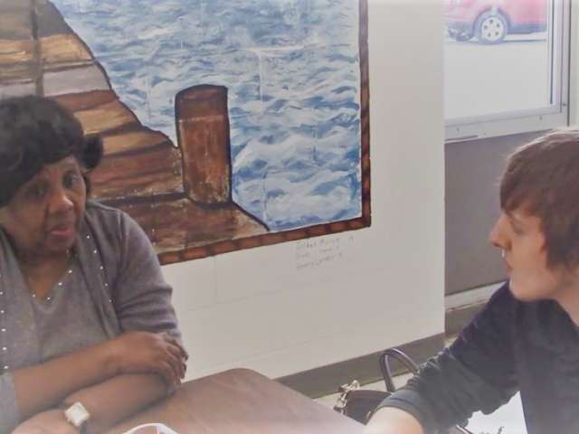 An African American woman sits with a white student in a classroom environment and chats with him.