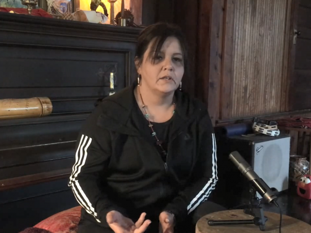 A woman with brown hair and a black running jacket sits in front of a microphone in her home. 