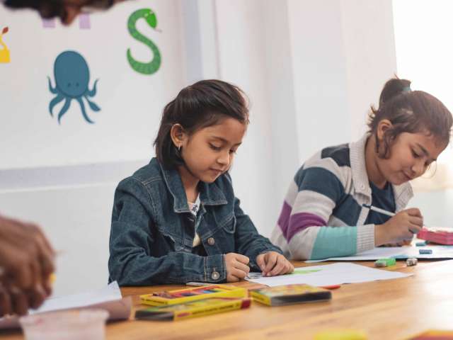 Two girls sit next to each in an elementary school classroom. 