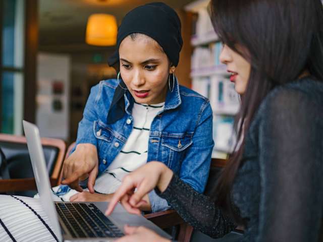 Two women sit in a communal space and look at a computer.