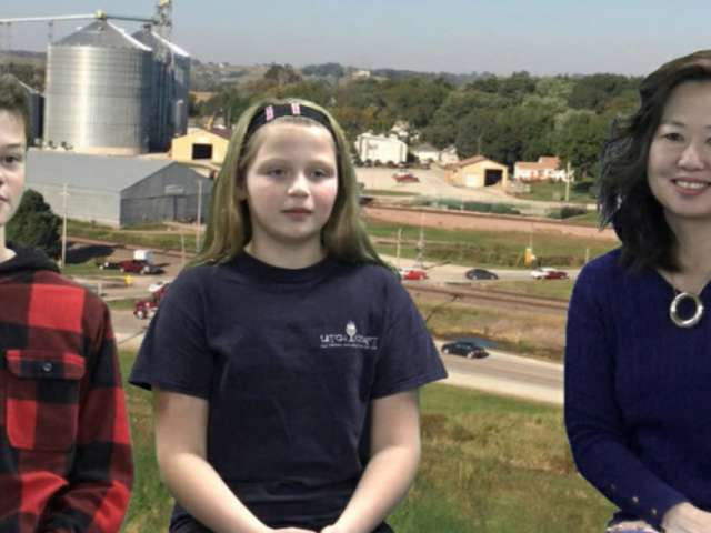 A young boy and girl sit with a woman in front of a farmscape as they talk to the camera. 