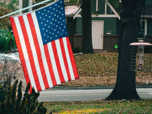 An American flag flies in a neighborhood in front of a green, cottage-style house. 