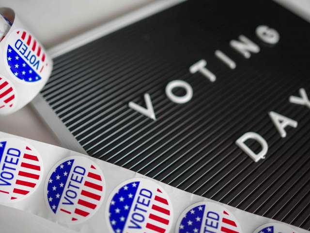 A black board shows the words, Voting Day, in a white font. The board is surrounded by round, I VOTED, stickers. 