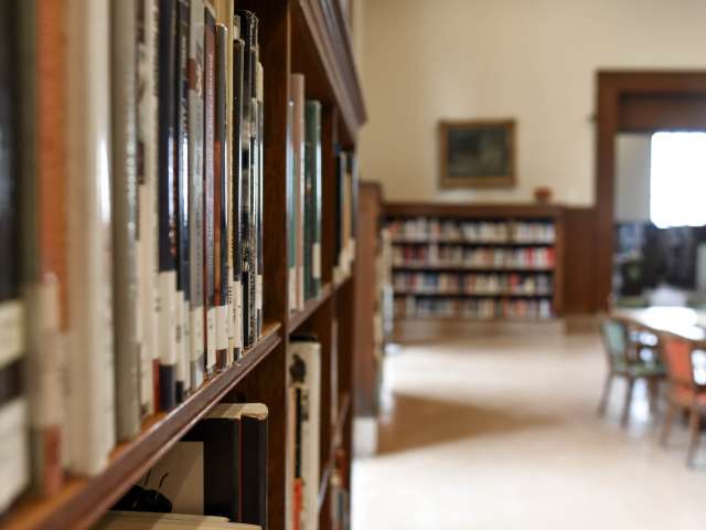The interior of a small school library, with a close-up of books on the shelf and desks and chairs in the background.