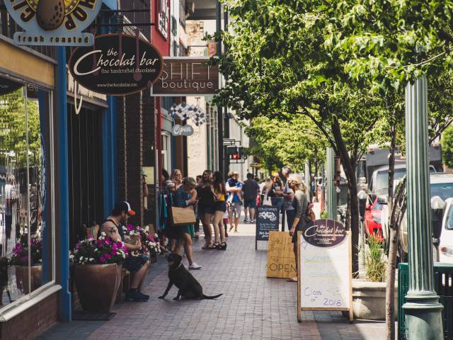 A small-town sidewalk on a summer day shows outdoor cafe tables and sign boards advertising sales. 