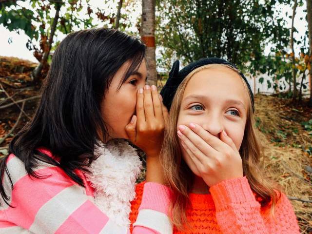 Two young girls in an outdoor setting whisper to each other and laugh.