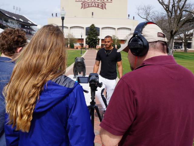 Three students, carrying video recording equipment, interview a young athlete on the campus of a college.