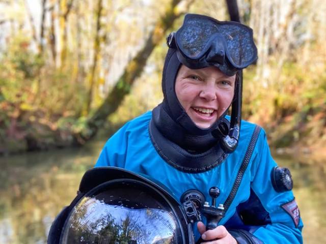 A woman wearing a blue dive suit and holding an underwater camera sits by a lake.