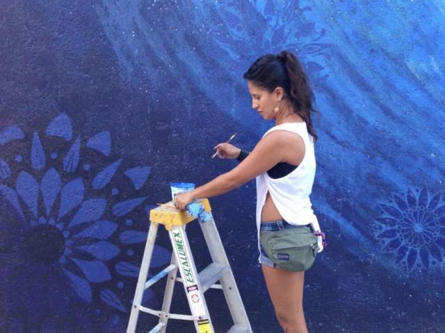 Adriana wears a white tank top and shorts as she stands on a ladder to paint a mural of water.