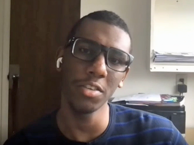Amir has short cropped hair, a blue t-shirt, and glasses. He sits in a dorm room.