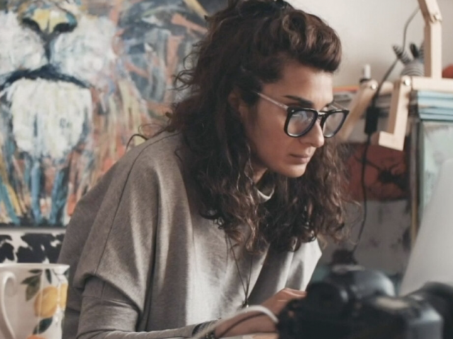 Asher has shoulder-length brown hair and thick reading glasses on. She sits at a desk with painting supplies.