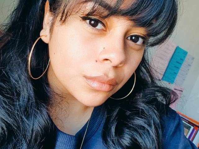 Arianna wears large, silver hoop earrings and has long, black hair as she sits in a classroom setting.