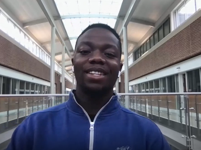Augustus wears a bright blue zip-up sweatshirt and sits in an academic building with classrooms in the background.