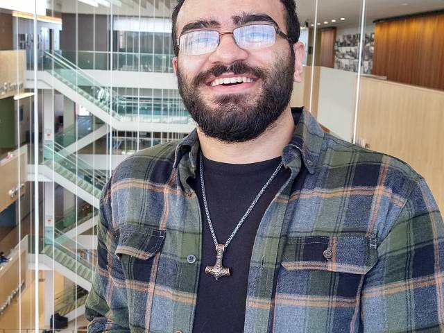 Balil has dark hair, a beard, and eyeglasses. He wears a blue, paid shirt in a library setting.