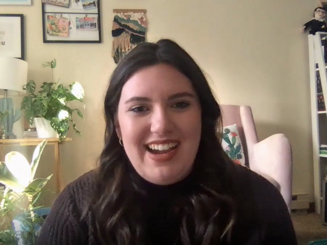Delanie has long, wavy brown hair and wears a black turtleneck sweater.