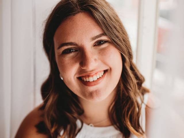 Delanie has long, wavy brown hair and wears a white tank top. She sits in front of a window with white curtains.