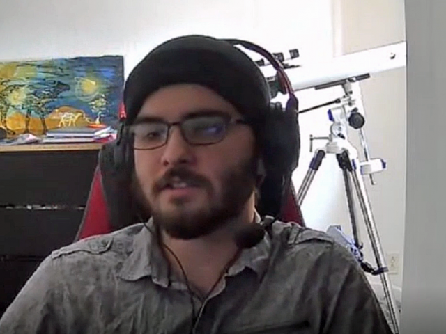 Dylan has a black knit cap on and headphones. He sits in front of a telescope and colorful painting.