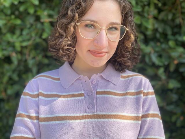 Sabrina has short, brown curly hair, eye glasses, and a purple and brown striped sweater on. She stands in front of greenery outside.