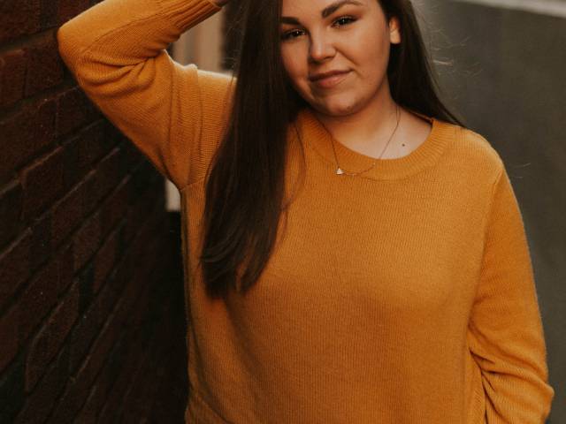 Sophia stands against the wall and puts her hand on her head. She wears a bright, orange sweater.