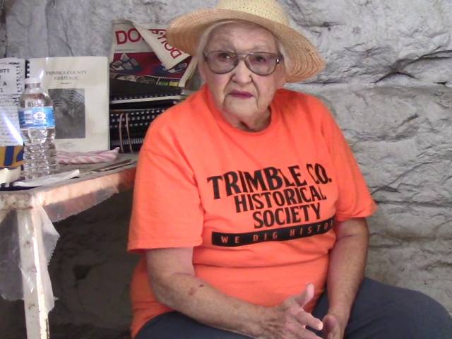A woman wears a straw hat and an orange shirt that says Trimble Co. Historical Society.