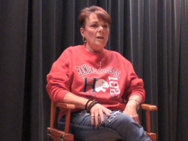 A woman with a red sweatshirt on sits in a director chair on a stage.