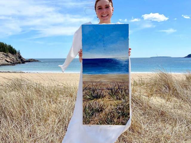 A woman stands on a beach and holds a painting up that matches the scenery exactly.