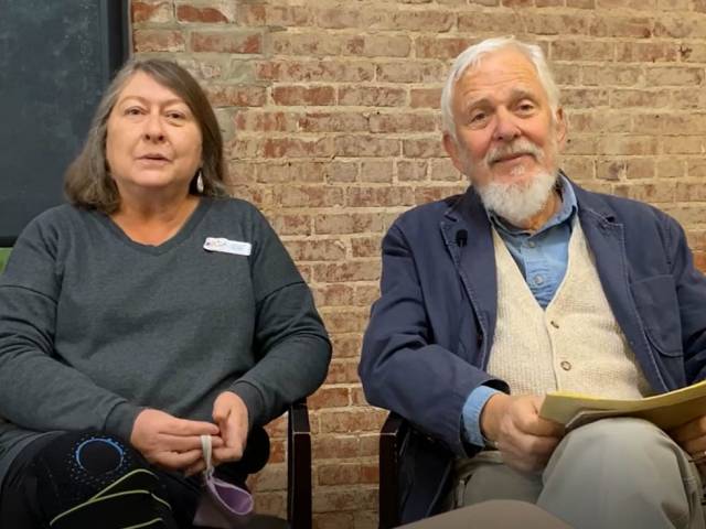 A man with white hair and a beard and a woman with shoulder-length brown hair sit in a library setting and chat with an interviewer. 