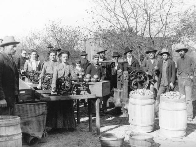 Vintage image of people in farm clothing standing around a market where foods and other goods are on display or for sale..
