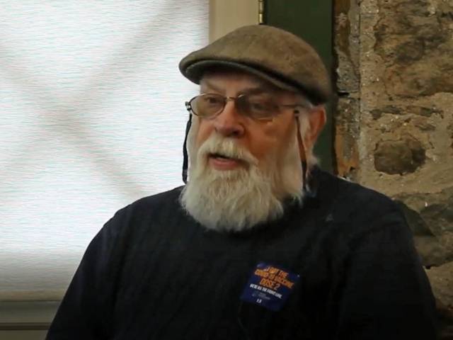 A man with a gray beard and a newsboy hat sits in front of a stone wall and chats with an interviewer. 