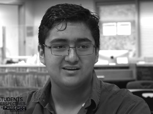 A young man with short, black hair and wire frame glasses sits in a library setting and is interviewed.
