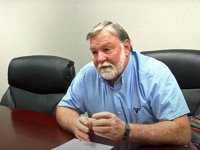 A man with gray hair, a beard, and a blue collared shirt sits in a black leather chair for an interview.