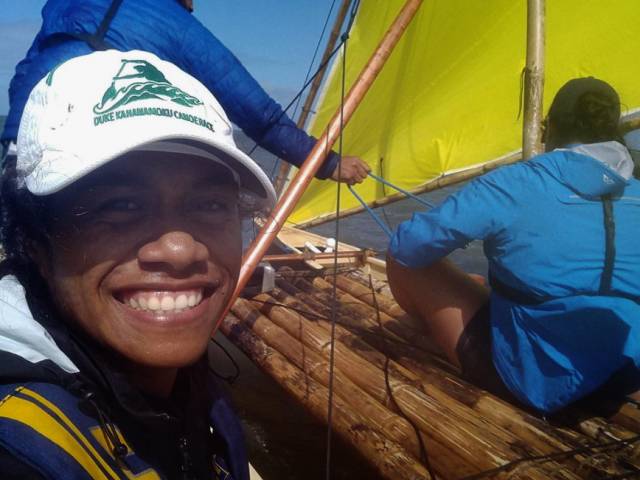 Suzanne wears a white baseball cap and sits on an outrigger on the water with a bright yellow sail.