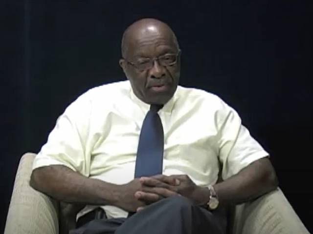 A man with a white-collared shirt and blue tie sits on a stage in a chair. 