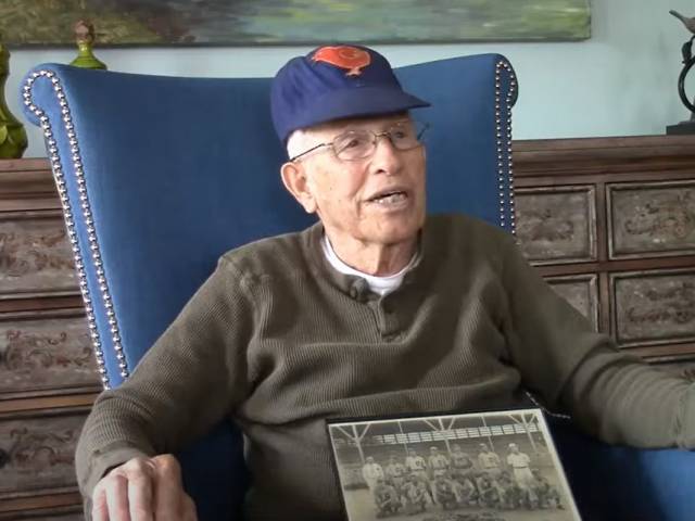 An elderly man with a baseball cap sits in a blue chair with an old photo on his lap.