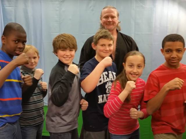 Children stand around a karate instructor and make a fist in support. 