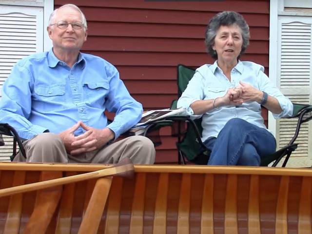 Two people sit outside on a porch with a canoe positioned in front of them.