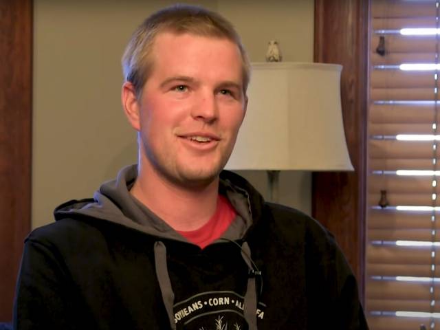 A young man with short blonde hair and a black sweatshirt sits on a couch with a window behind him.