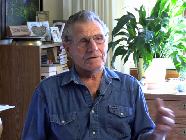 A man with gray hair and denim shirt sits in a living room with a plant and window behind him.