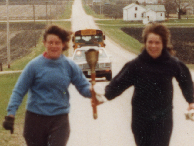 Two women running along a rural road carrying an Olympic torch.