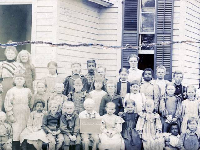 A large group of children stand in front of an old school house around the turn of the 20th century.