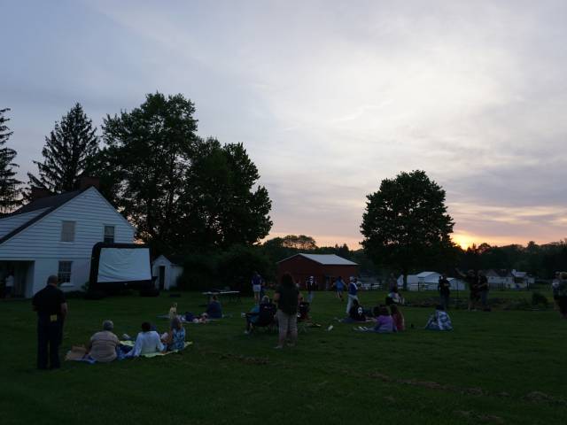 A white farm building and people gathered on the lawn of it at dusk.