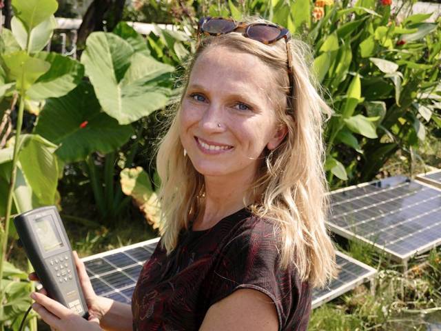 A woman with a black shirt and sunglasses on her head stands in a garden. 