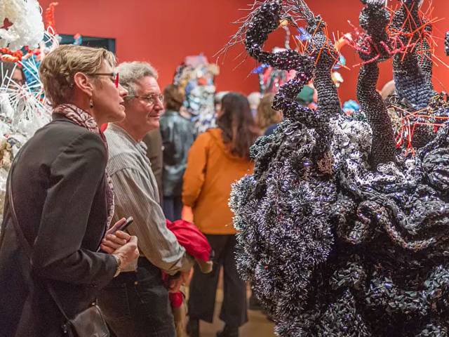 Two women look at a crocheted coral reef in a red gallery space. 