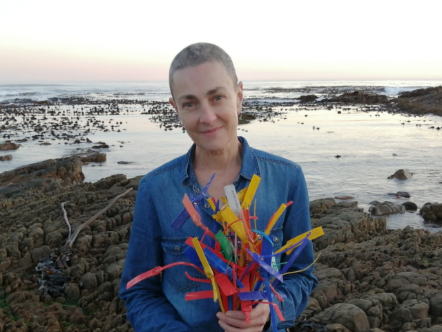 A person stands next to a rocky shore. She wears a blue blazer and hold a colorful plastic sculpture.