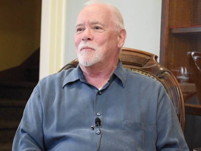 A man with a white beard wears a button-down blue shirt and sits in a vintage chair for an interview.