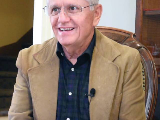 A man with white hair and a suede brown jacket sits in a vintage chair. 