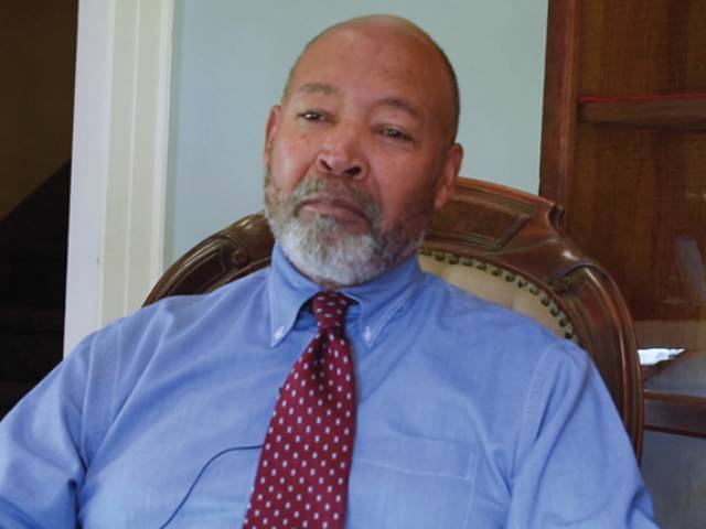 A man with a gray beard and blue collared shirt and red tie sits in a vintage style chair.