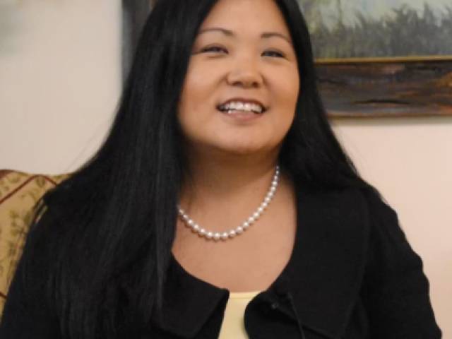A woman with long black hair, a black sweater and a pearl necklace sits for an interview.
